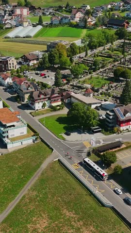 Vertical aerial view over Schwyz, Switzerland, with cyclists and a public bus crossing a town junction beside homes, greenhouses and cultivated fields on a sunny summer day.