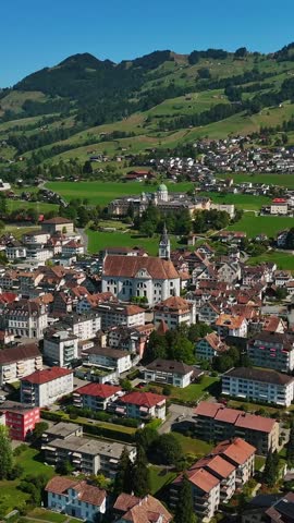 Vertical aerial panorama over Schwyz, Switzerland, with St. Martin Church dominating the historic center, surrounded by red roofs, green fields and steep Alpine hills.