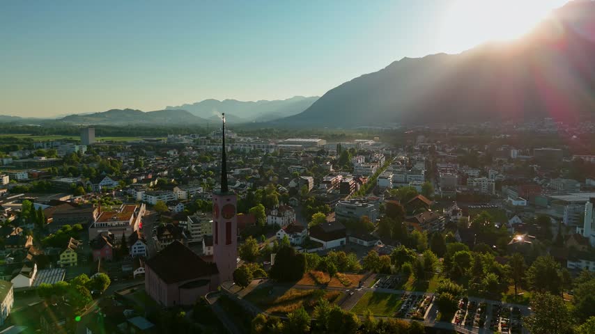 Golden-hour aerial view over Buchs SG, Switzerland, with the Evangelische Kirche Buchs, dense neighborhoods and dramatic Alpine mountains glowing beneath the setting sun.
