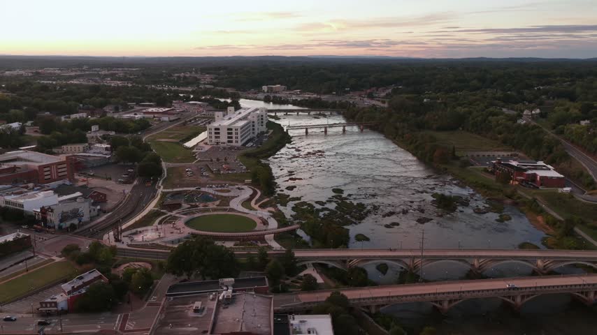 A multi arch concrete bridge spans the Dan River at dusk in Danville, Virginia, a white riverside building glowing pink beside the rippled shallows below. Aerial view