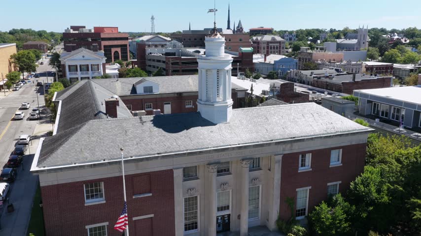 A red brick civic building with a white cupola and half staff flag stands in downtown Wilmington, North Carolina, church spires and rooftops spreading behind it. Aerial view