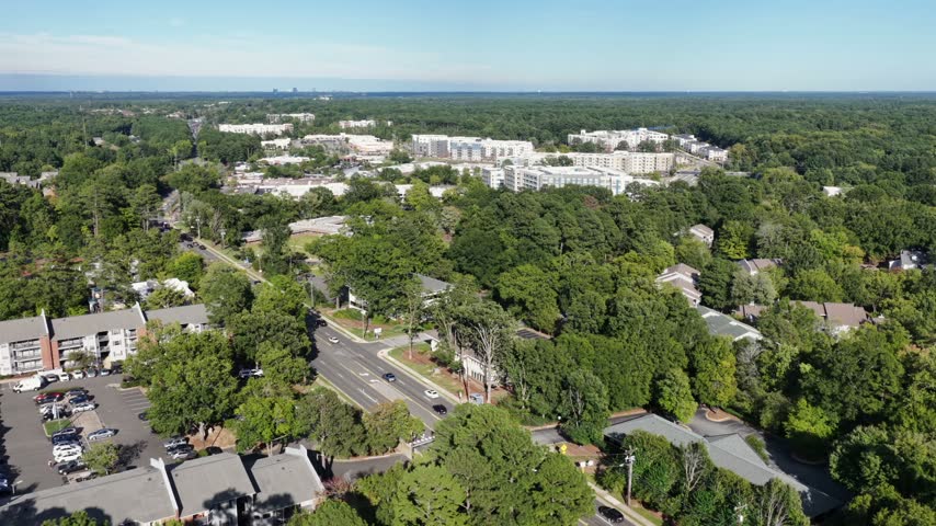 Tree shaded apartment buildings with a swimming pool spread through the woods near Duke University in Durham, North Carolina, parking lots visible beside a busy road. Aerial view