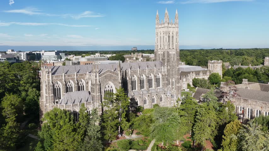 A close orbit of Duke Chapel shows its buttressed nave and arched windows in Durham, North Carolina, campus paths and lawns spreading beneath the tower. Aerial view