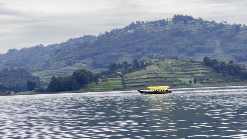 Covered passenger motorboat travels across Lake Bunyonyi, southwestern Uganda, with a bright yellow canopy, narrow hull and terraced Kigezi Highlands rising behind, wide telephoto view.