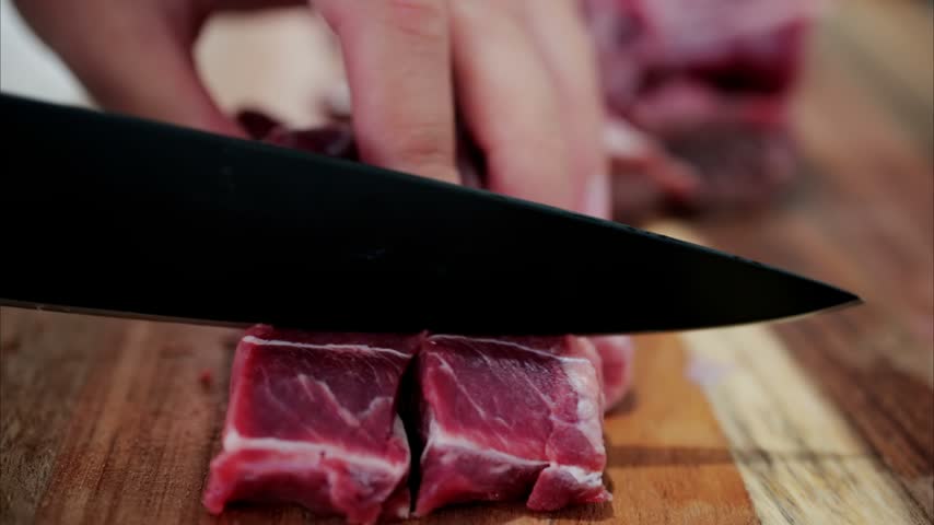 Professional chef cutting fresh raw steak into small cubes on wooden board in kitchen studio, culinary preparation process.