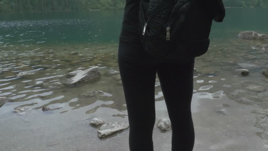 Hiker woman with a backpack walks to the transparent water of Morskie Oko lake and looks at the forested slopes of the Tatra Mountains in Tatra National Park.