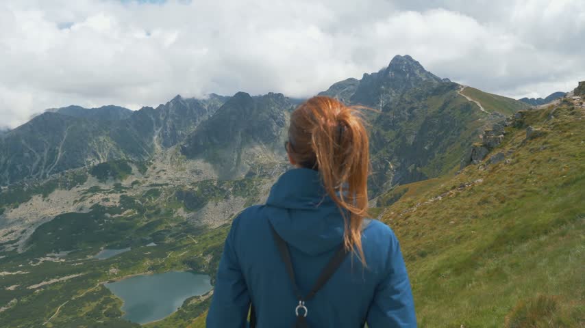 Woman in a blue jacket with windblown ponytail looks from a ridge over the Gasienicowa Valley, its glacial lakes and rocky peaks in the Tatra Mountains of Poland.