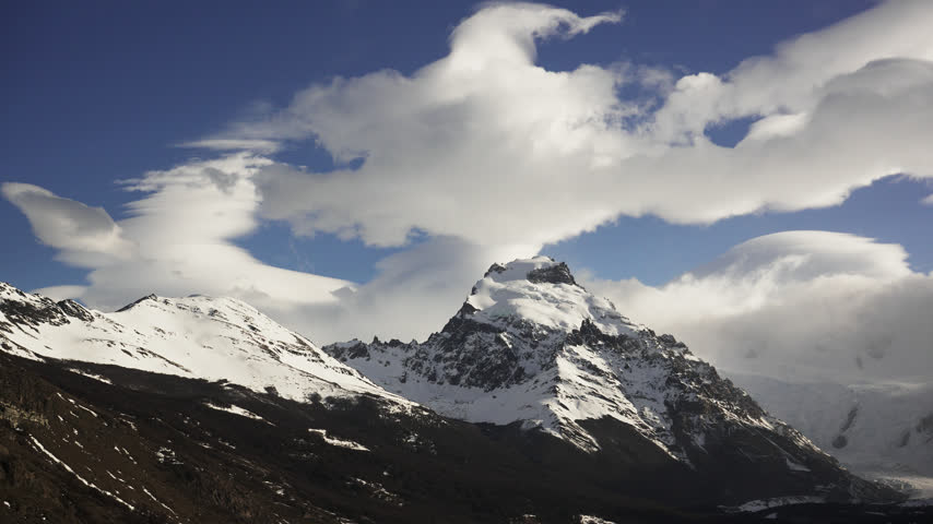A snow covered mountain rises above dark wooded slopes beneath sculpted white clouds and blue sky near El Chalten in Santa Cruz province in Argentine Patagonia