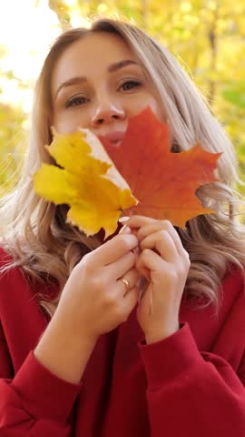 Woman playfully hides behind autumn leaves, holding orange and yellow foliage near her face. Smiling woman lowers foliage outdoors, enjoying bright fall colors while wearing red sweater.