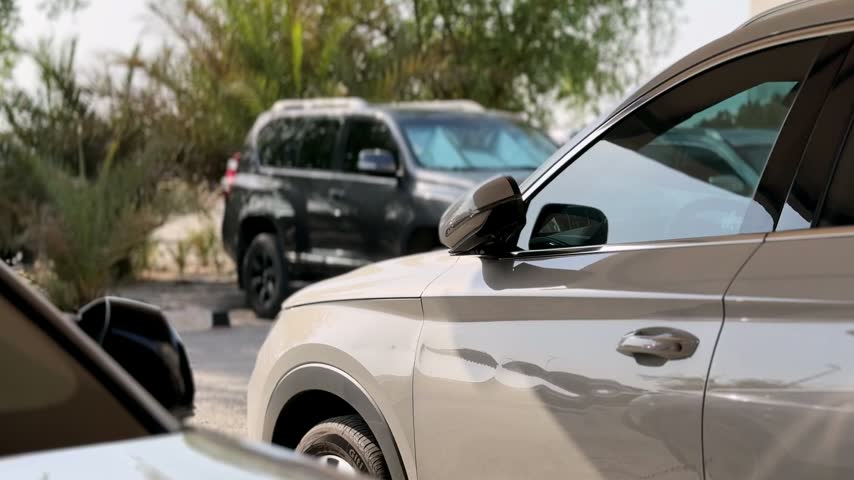 Close up of parked modern cars in an outdoor lot during bright daylight. Cinematic shallow depth of field highlights a light grey SUV side against trees and other vehicles.