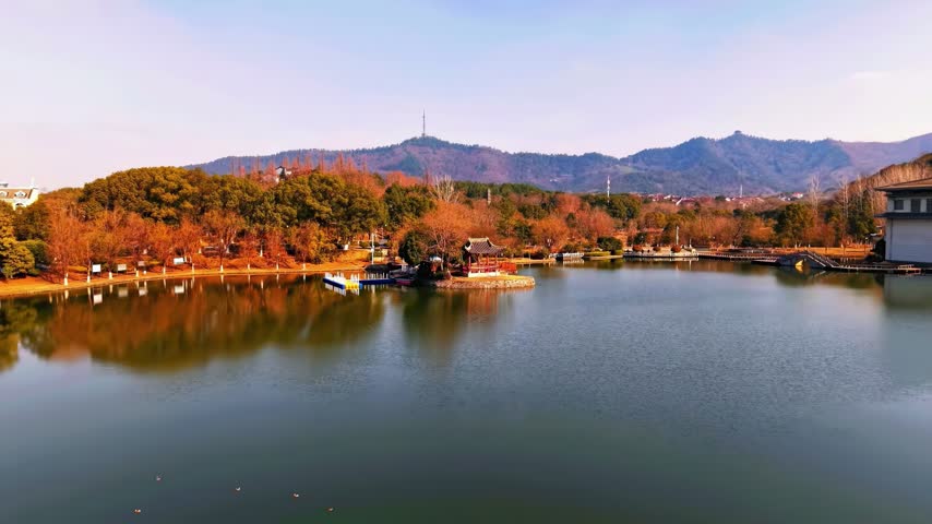 A serene traditional Chinese pavilion sits on a small island in the middle of a calm lake. The surrounding park is filled with autumn trees and distant mountains under a clear blue sky.