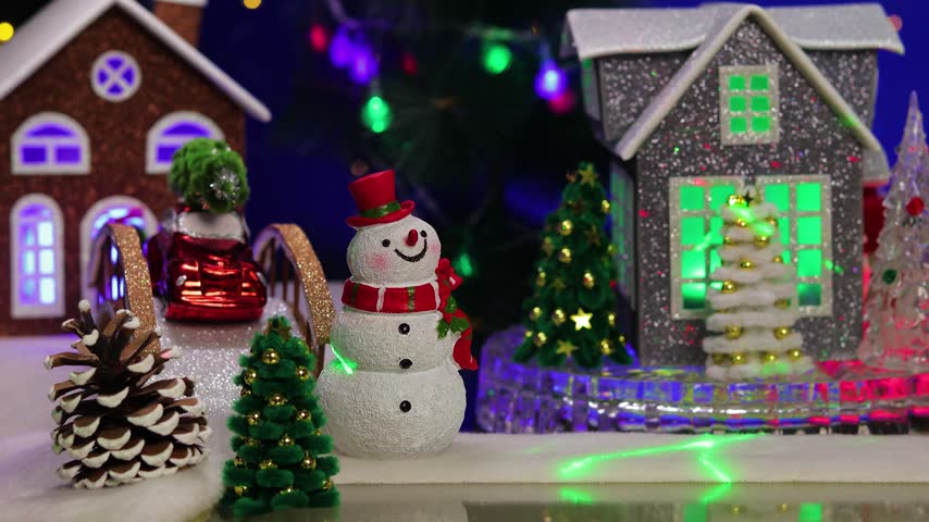 In the Christmas village, a grey house rotates, surrounded by small fir trees and snowmen