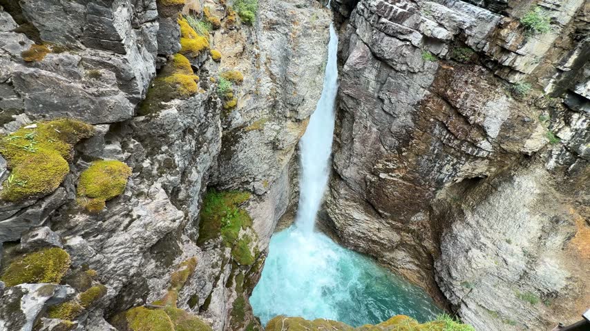 A waterfall plunges into a vivid turquoise pool deep in Johnston Canyon at Banff National Park, Alberta, surrounded by steep mossy limestone cliffs and pine forest.