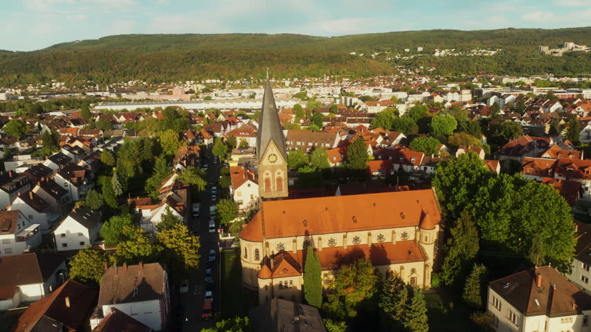 Munich church recedes as the drone pulls back over roofs beneath wooded hills.