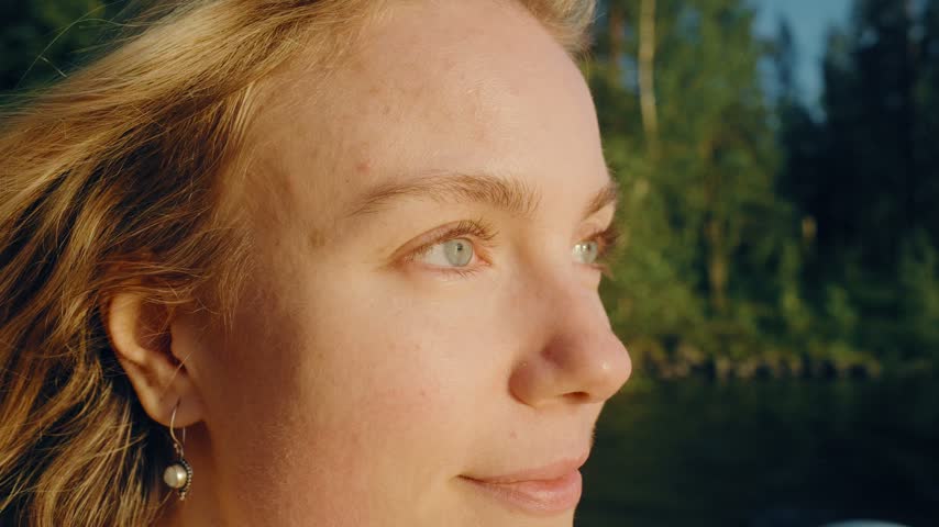 Close-up shot of a beautiful blonde woman with striking green eyes looking thoughtfully at a sunlit landscape, surrounded by lush greenery, in soft warm evening light.