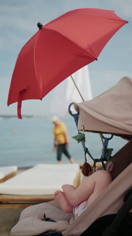 Baby sitting in stroller under bright red beach umbrella by the sea