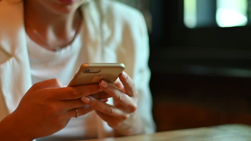 Young woman holding smartphone and scrolling through feed or messaging while sitting at indoor table, social media engagement.