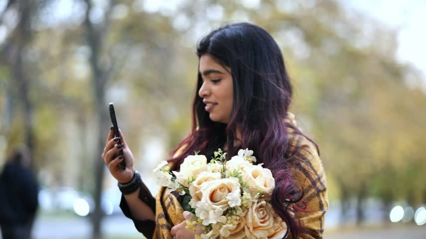 Young Indian woman holding flowers, smiling during a video call in autumn park