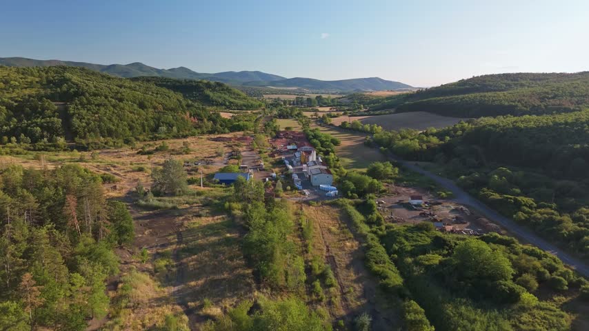 Aerial view of an industrial facility amid cultivated fields and forested hills in the Mátra Mountains near Rákóczibánya, Hungary, on a clear summer day.