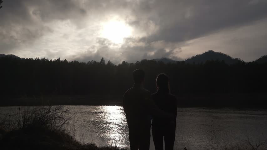 Dark silhouettes of a man and a woman embracing, standing on the riverbank with their backs to the camera at dusk after sunset. Love, intimacy, trust, expectation.