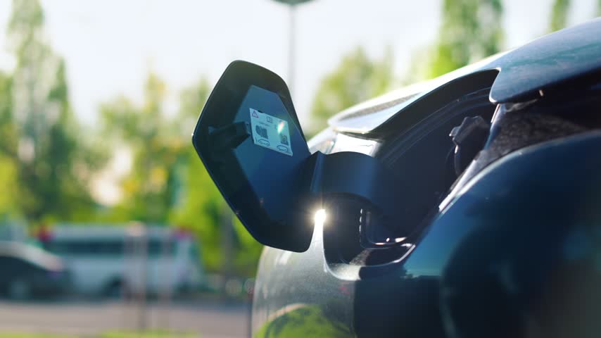 Close up of male hand plugging charger connector into electric car inlet with bright sun flare on background.