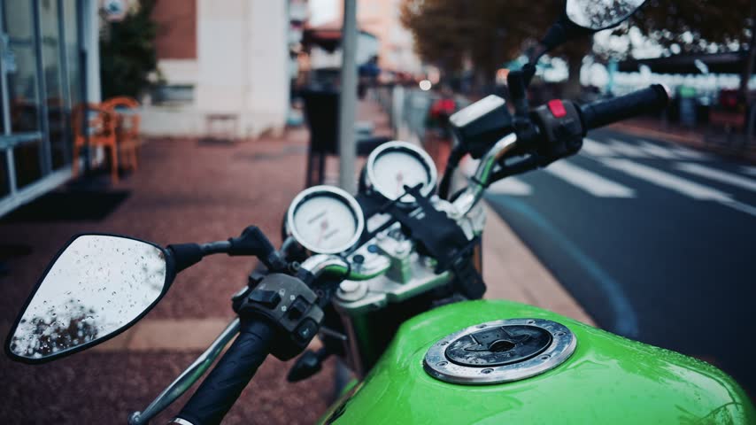 Close up of bright green motorbike cockpit covered in water droplets parked near road with blurred city background.