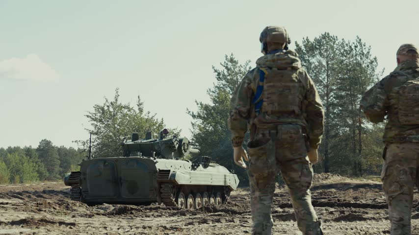 A soldier raises his hand to signal direction to an approaching armored tracked vehicle on a sandy training field.