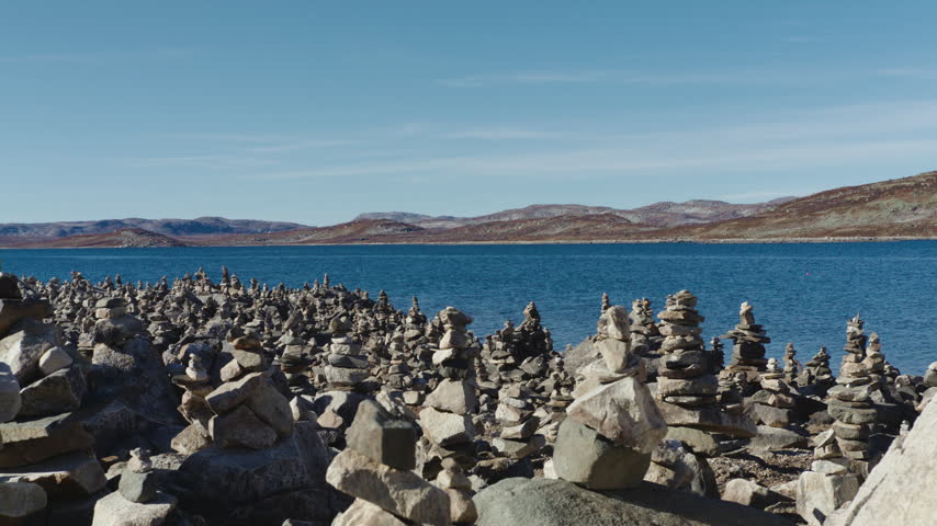 Crowded rock piles cover a barren shoreline along a deep blue highland lake under a clear sky, with low brown ridges stretching across the horizon.