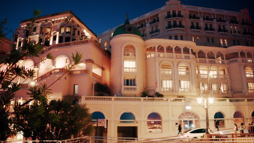 Elegant architecture illuminated by warm lights at night in Monaco with cars passing by on street, cinematic city scene.