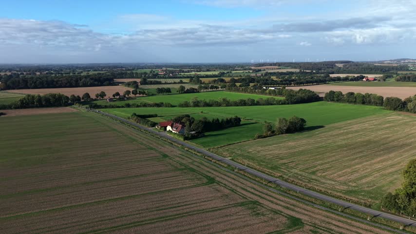 Red-roofed farmhouse sits amid patchwork green and golden harvested fields lining a quiet American country road, distant wind turbines under partly cloudy autumn sky. Aerial drone wide shot