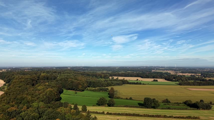 Quiet rural autumn landscape with muted green fields, dense forest patches, distant wind turbines under wispy blue sky over peaceful open countryside in United States. Aerial drone wide shot