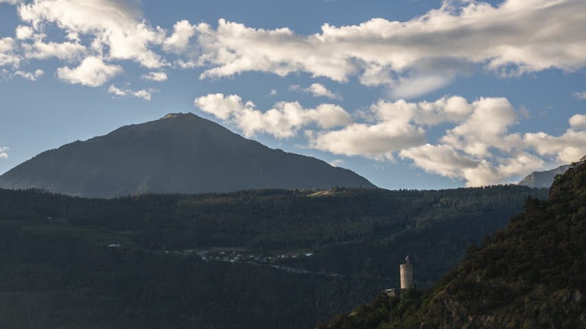 Timelapse of clouds crossing a forested mountain valley above Martigny, Switzerland, with a round stone tower and Swiss flag overlooking the alpine landscape.