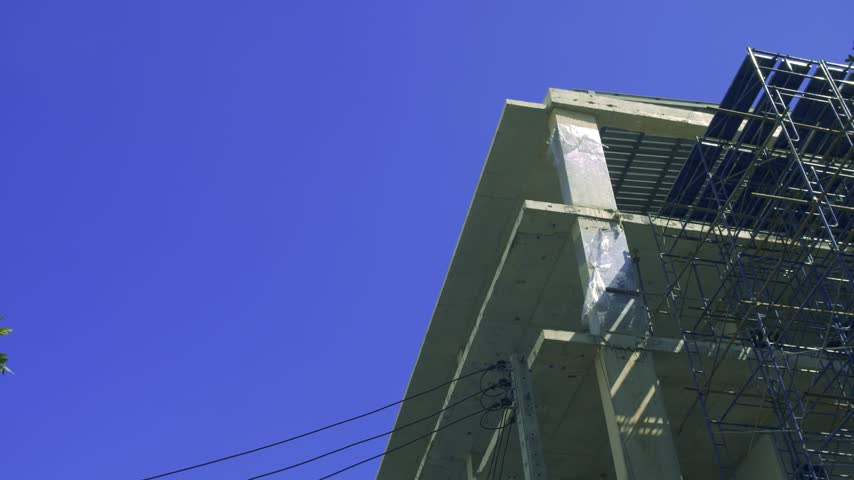A tilting up shot of a concrete building corner wrapped in blue scaffolding against the sky