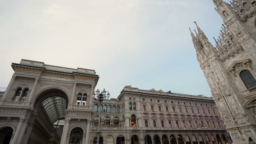 Low angle point of view shot walking in piazza del duomo, revealing the magnificent gothic facade of the milan cathedral and the galleria vittorio emanuele ii on a cloudy summer evening
