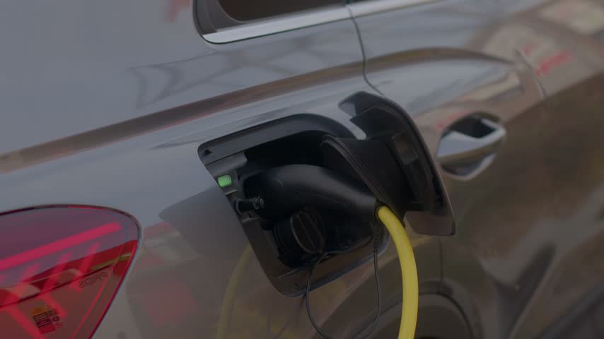 Close-up view of electric car charging port with bright yellow cable plugged in, green LED indicator lit, metallic bodywork and rear taillight visible in outdoor daylight.