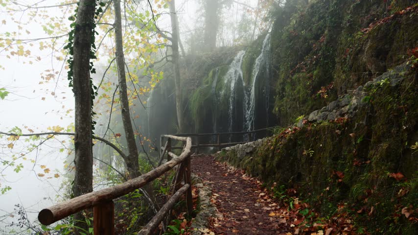 Scenic stone trail with wooden railing next to cascading waterfall in damp foggy forest during autumn season.
