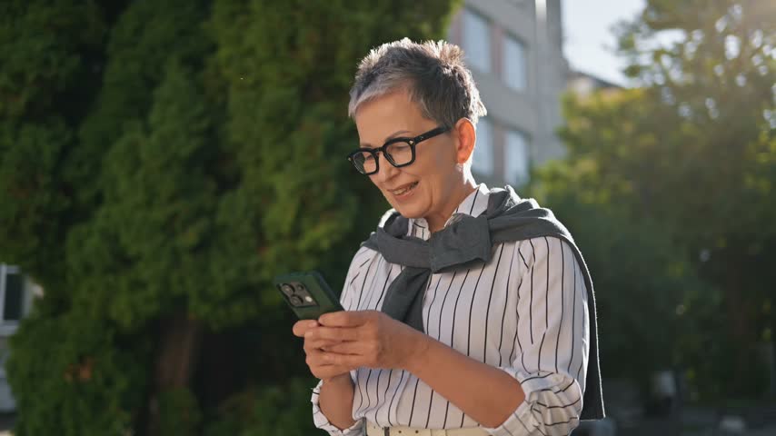 Older woman with glasses checks her smartphone in a park, surrounded by greenery and benches, enjoying a sunny day outdoors