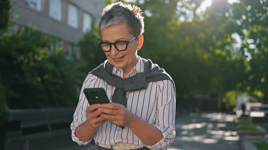 Older woman with glasses checks her smartphone in a park, surrounded by greenery and benches, enjoying a sunny day outdoors