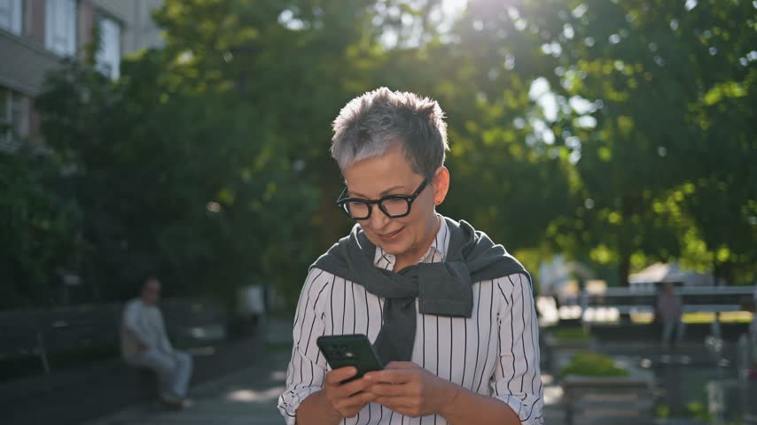 Older woman with glasses checks her smartphone in a park, surrounded by greenery and benches, enjoying a sunny day outdoors