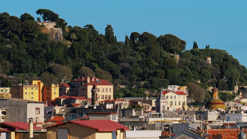 Scenic view featuring majestic waterfall flowing on tree covered hill above historic colorful old town rooftops in Nice France.