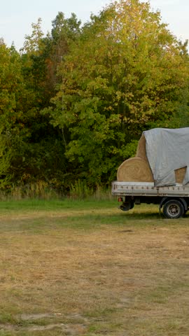 Parked flatbed truck sits on grassy field, carrying large round hay bales covered with weathered tarp, with nearby trees starting autumn colors.