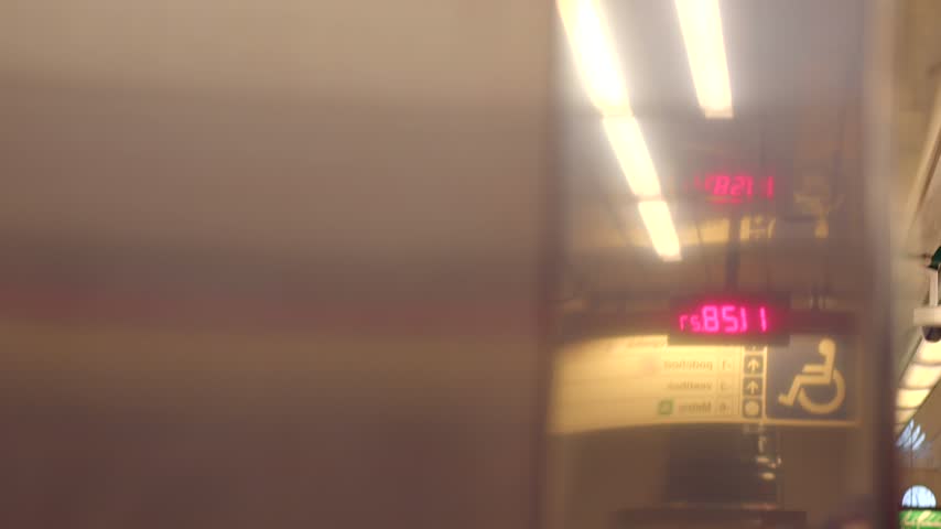 Reflective metal panel inside subway station with warm overhead lights, red digital clock, wayfinding signage, and visible platform edge.