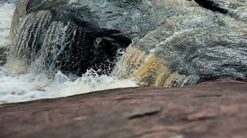 Closeup view of fast flowing stream water cascading over smooth dark rocks and creating foamy white rapids