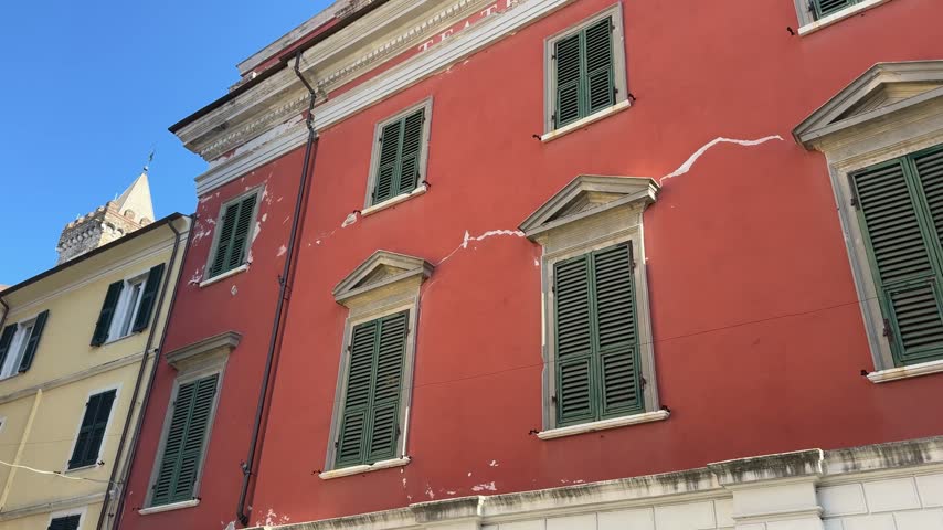 A low-angle view of vibrant red and yellow historic buildings with green shutters and weathered facades in the old town of Sarzana, Liguria, under a clear blue sky.