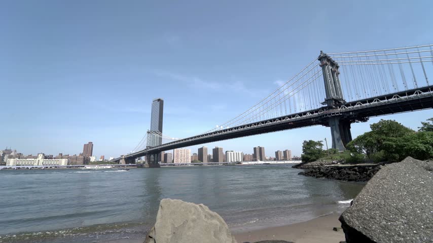 Small waves wash onto a sandy shore beneath a steel suspension bridge on the Brooklyn waterfront, with rocks in the foreground and Manhattan across the East River in New York City.