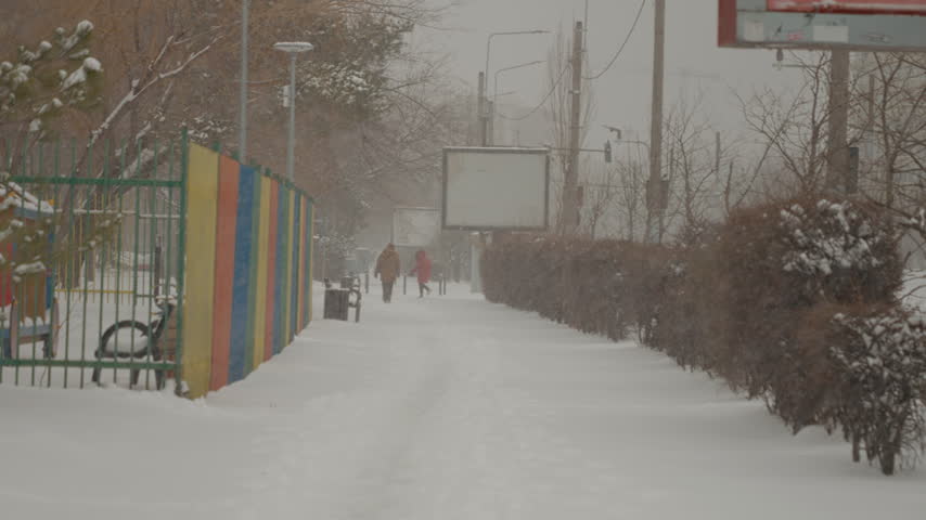 Snow falls steadily over city streets, creating a winter wonderland. Pedestrians walk through the snowfall, wrapped up warm against the cold.