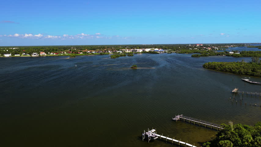 An aerial view moves toward a small vegetated islet across rippling water, with docks and houses along the distant shore.