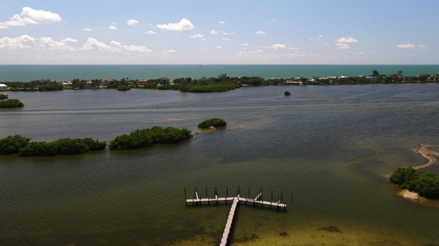 An aerial view shows a dock extending into a lagoon beside mangrove islets, with waterfront houses and the open sea beyond.