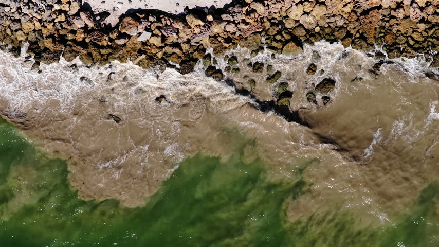 An overhead view shows waves washing over shoreline rocks and sand, with brown sediment spreading into green seawater.