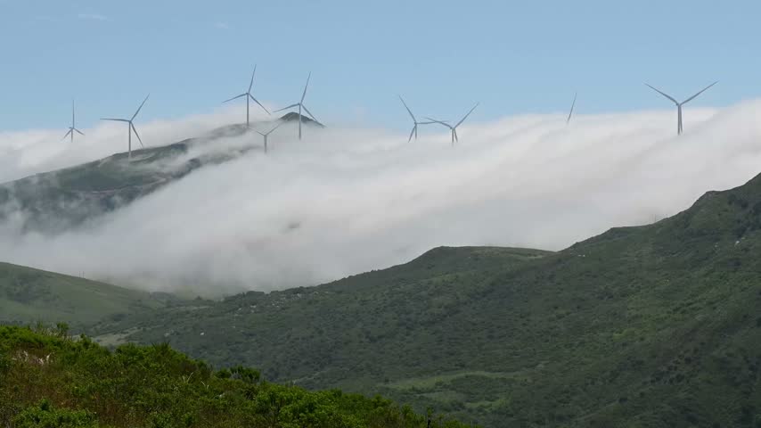 Scenic landscape featuring green grassy hills and rolling mountain peaks partially covered by a low layer of clouds or fog against a blue sky.